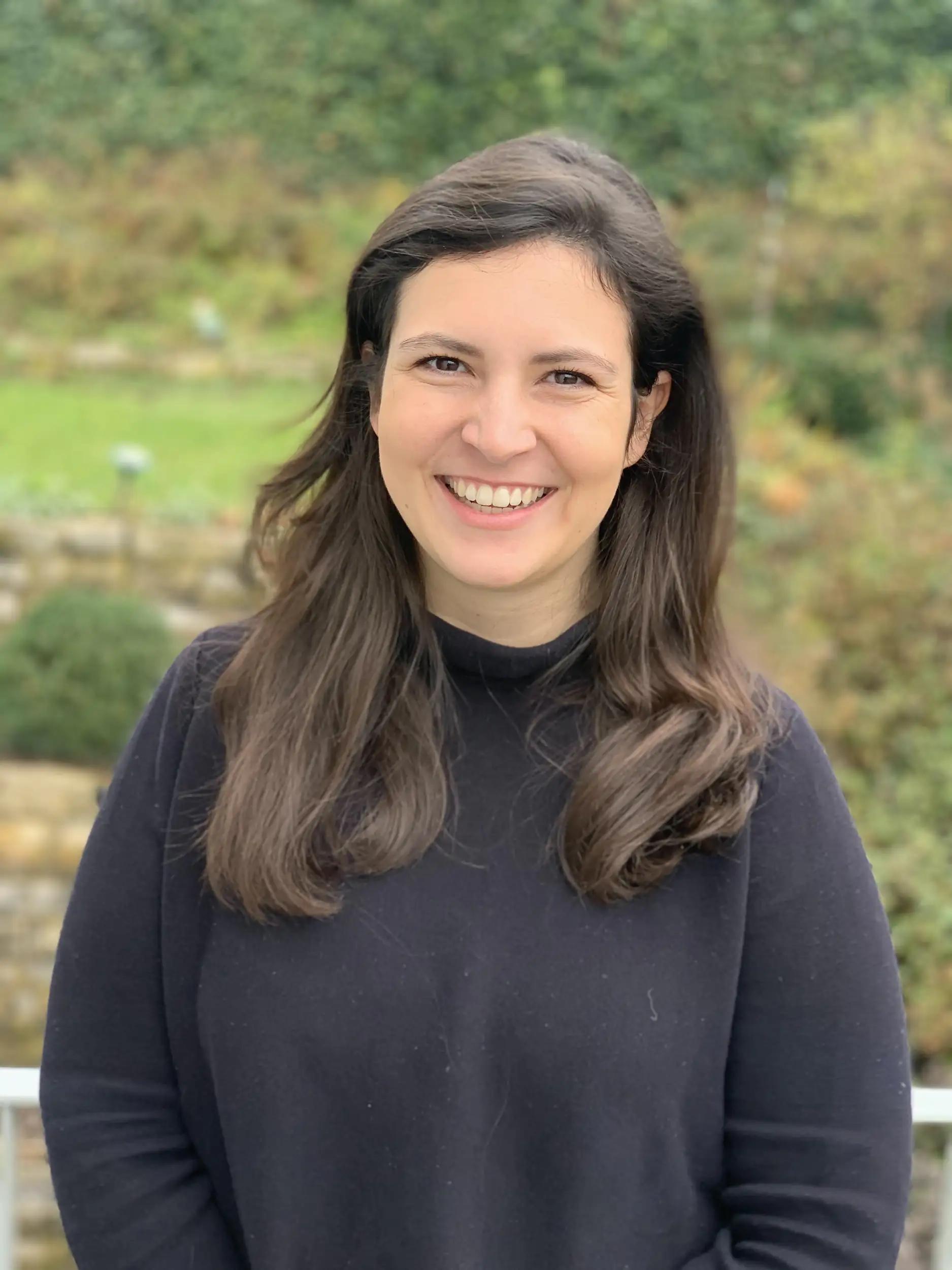 Woman with long brown hair smiling outdoors wearing a black sweater.