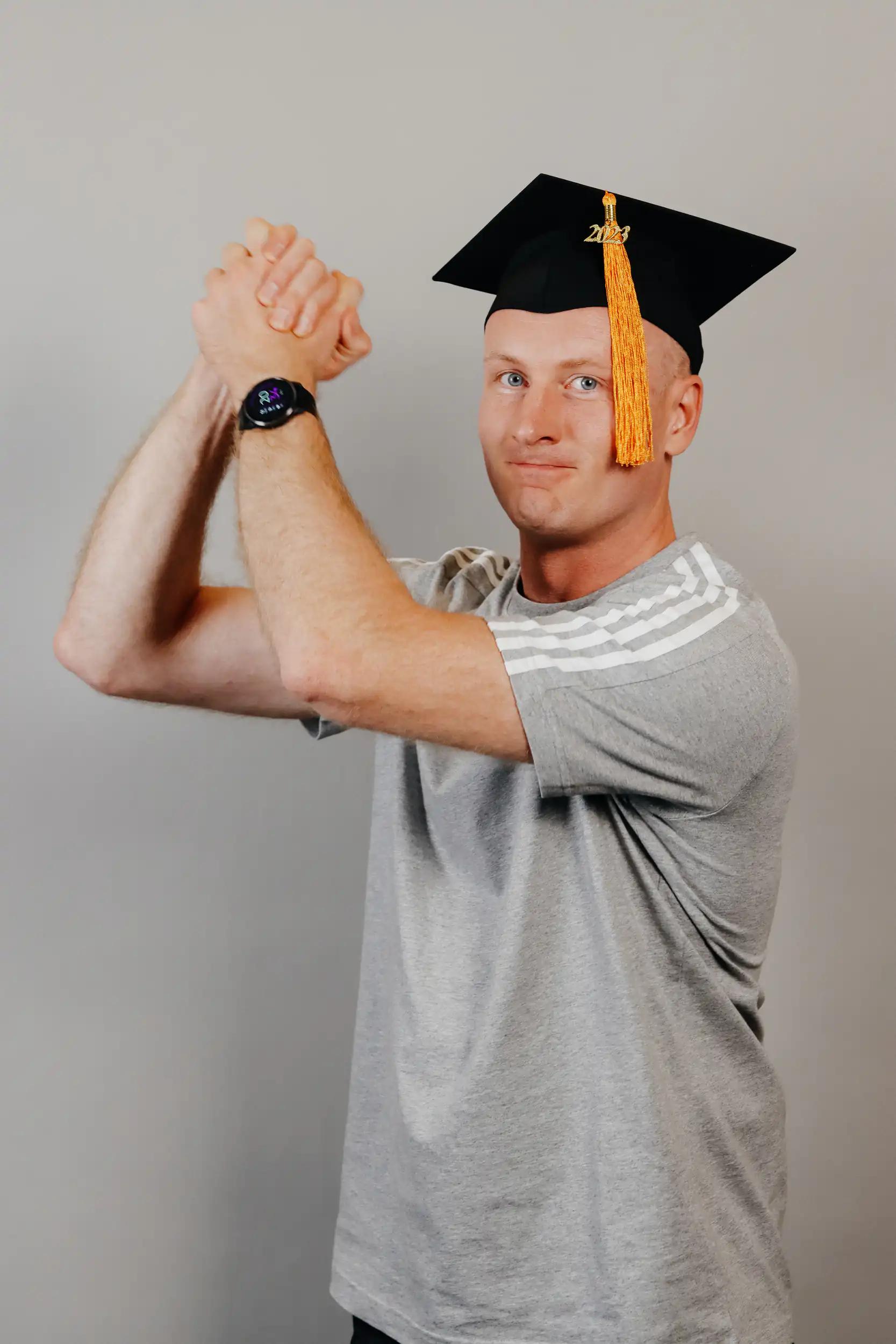 Mann mit einem grauen T-Shirt und einem schwarzen Absolventenhut mit Quaste posiert.