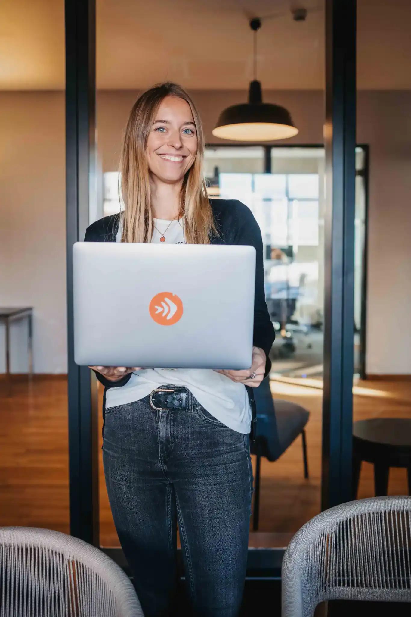 Frau hält einen Laptop und lächelt in einem Büro.