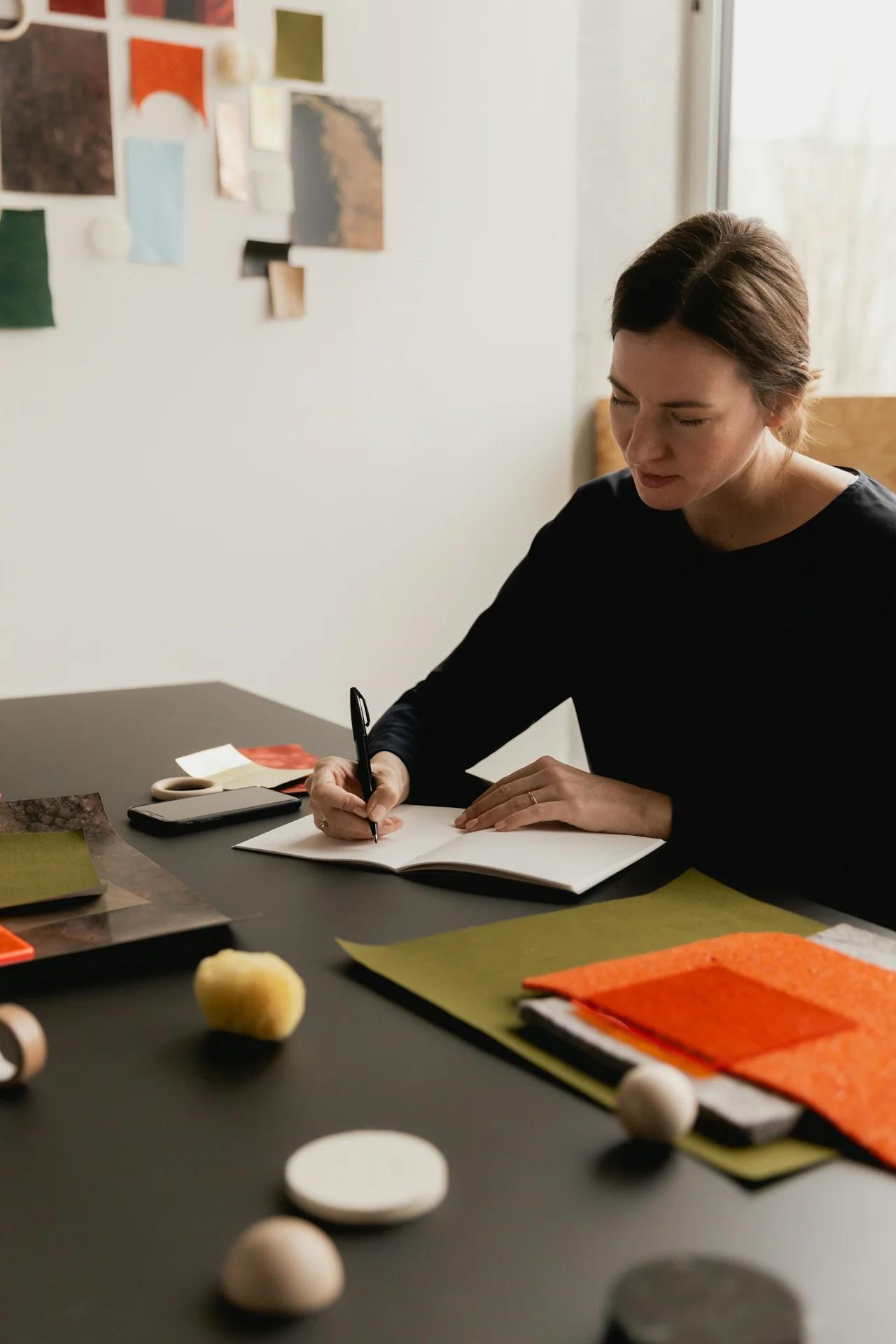 Frau schreibt in Notizbuch an Tisch mit Designmaterialien.