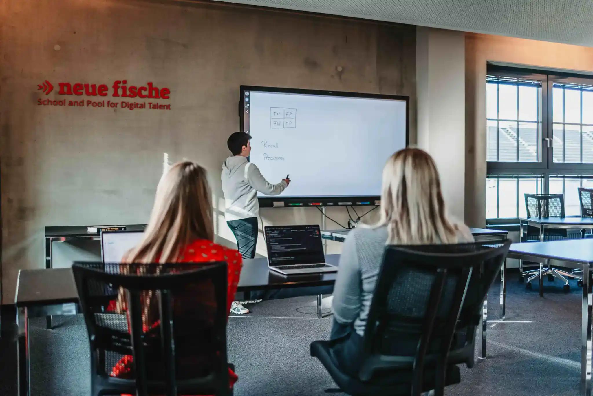 Mann erklärt etwas an einem digitalen Whiteboard vor zwei Personen im Büro.