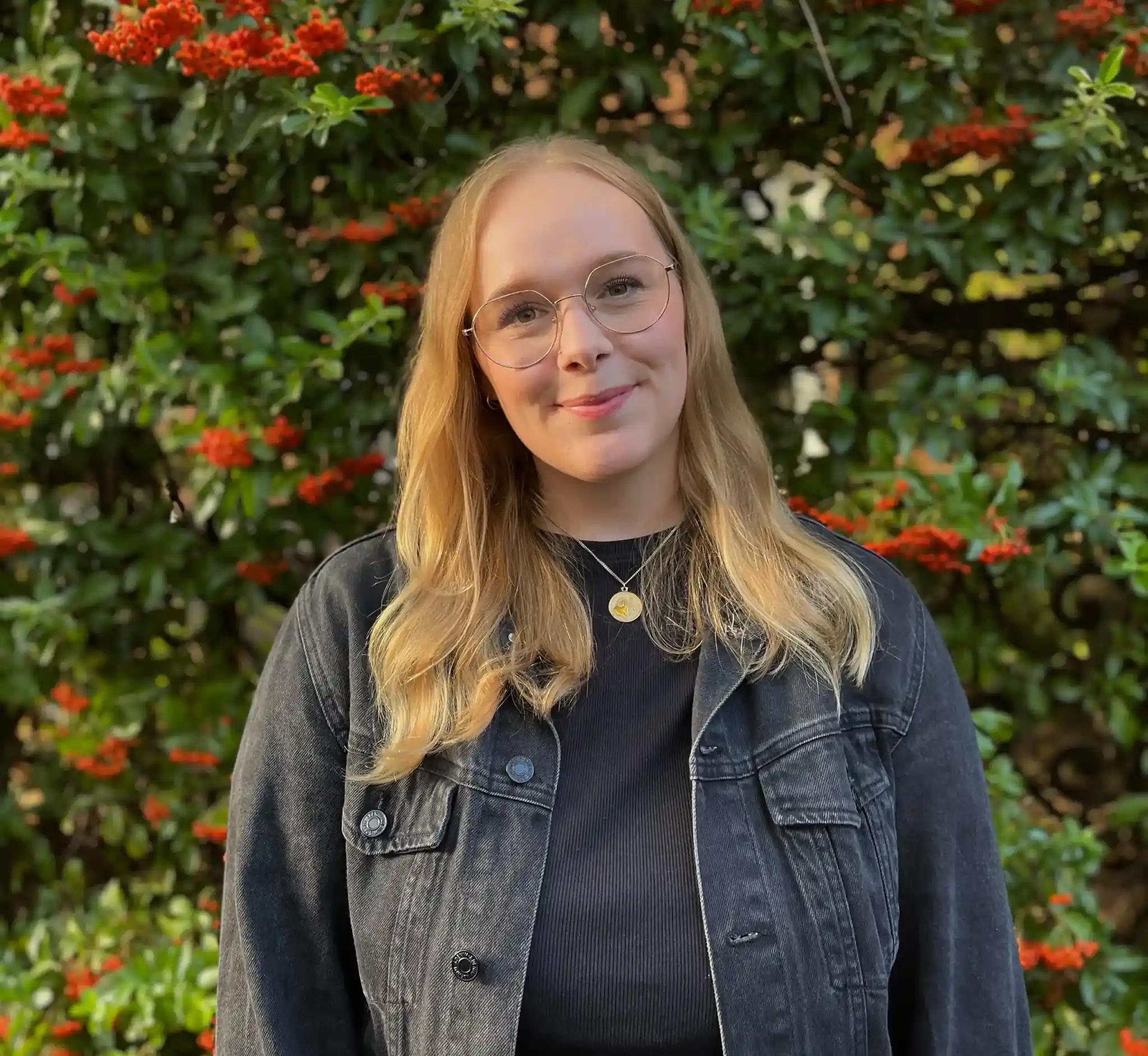Porträt einer jungen Frau mit Brille vor einer Pflanzenwand mit roten Beeren.