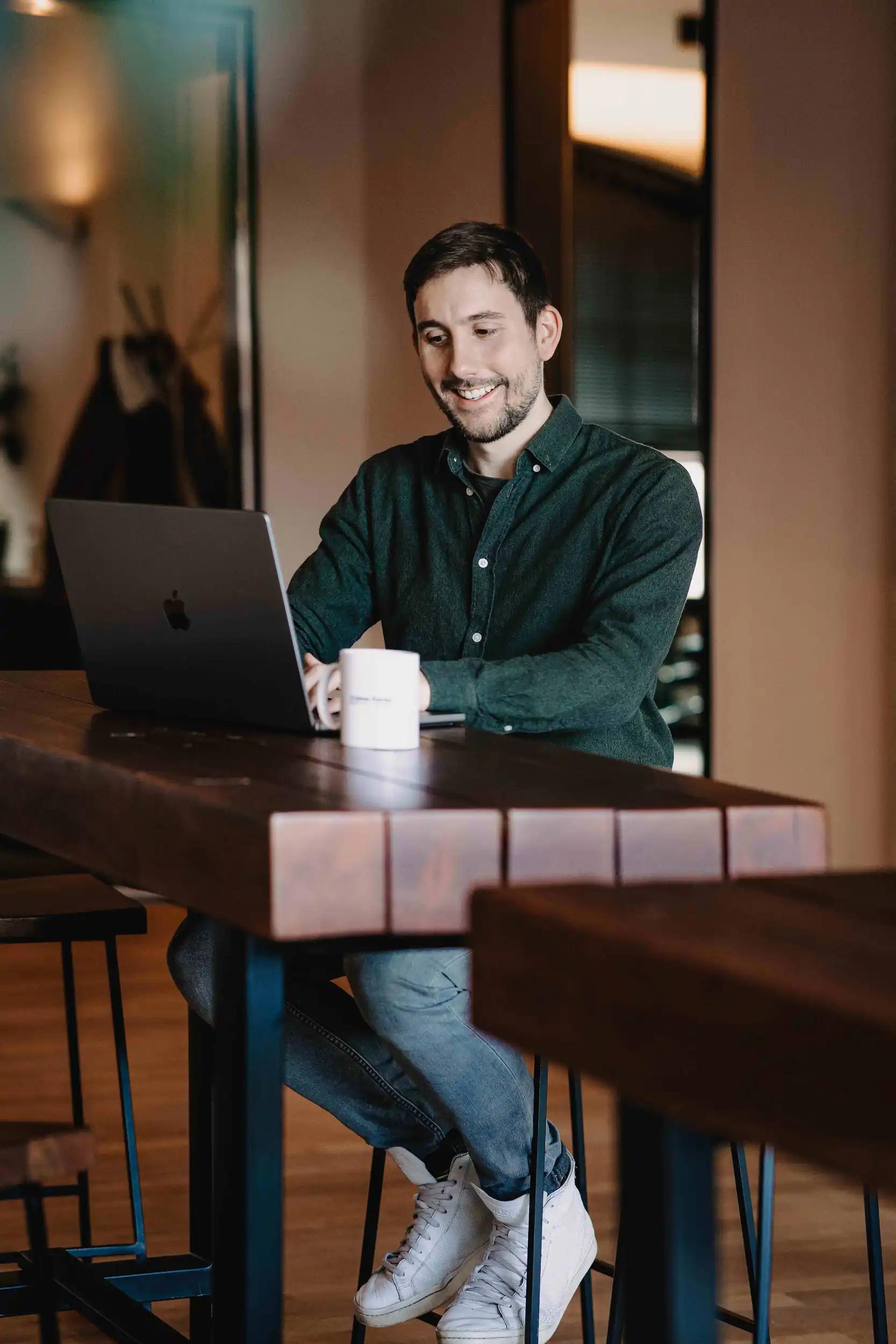 Man sitting at a wooden table working on a laptop and smiling.