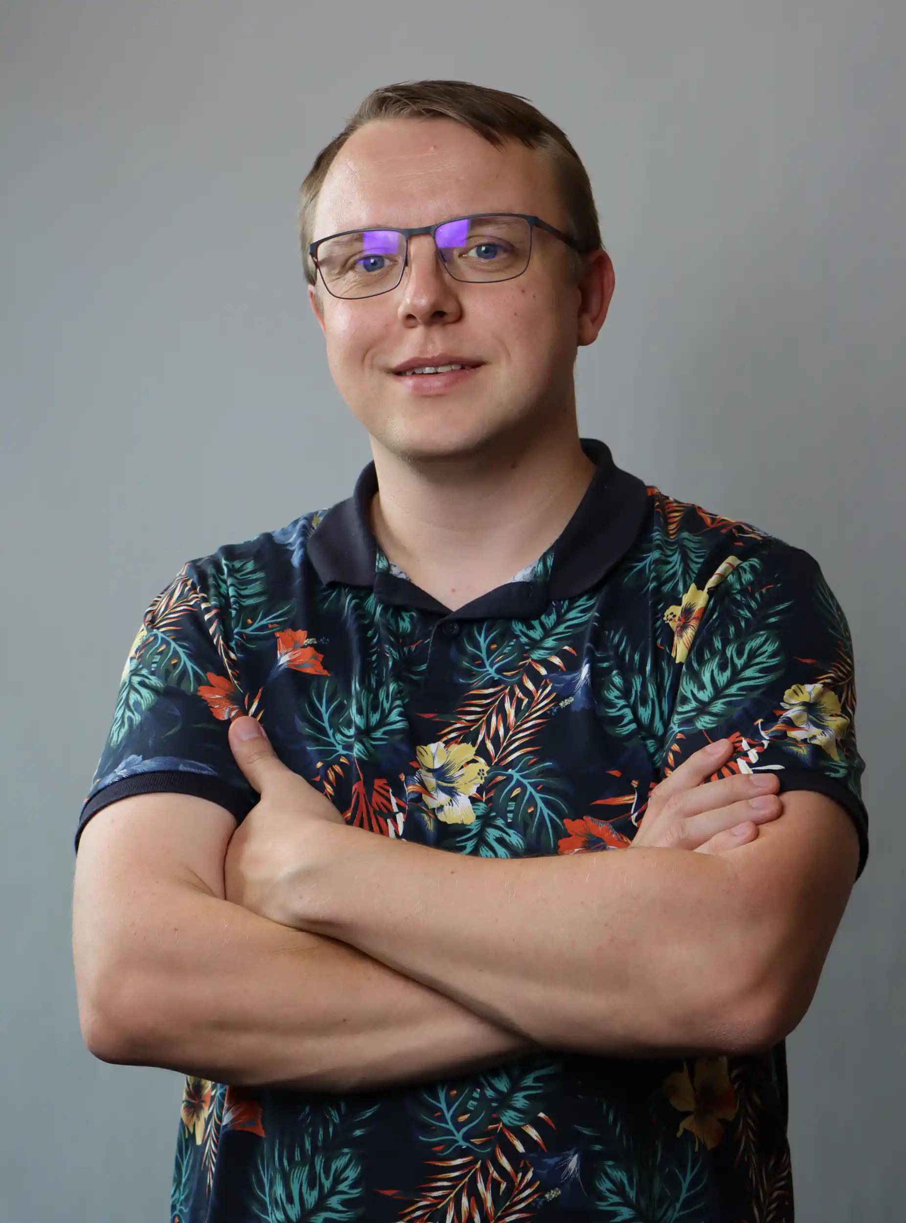 Mann mit Brille und floralem Hemd mit verschränkten Armen vor grauem Hintergrund.