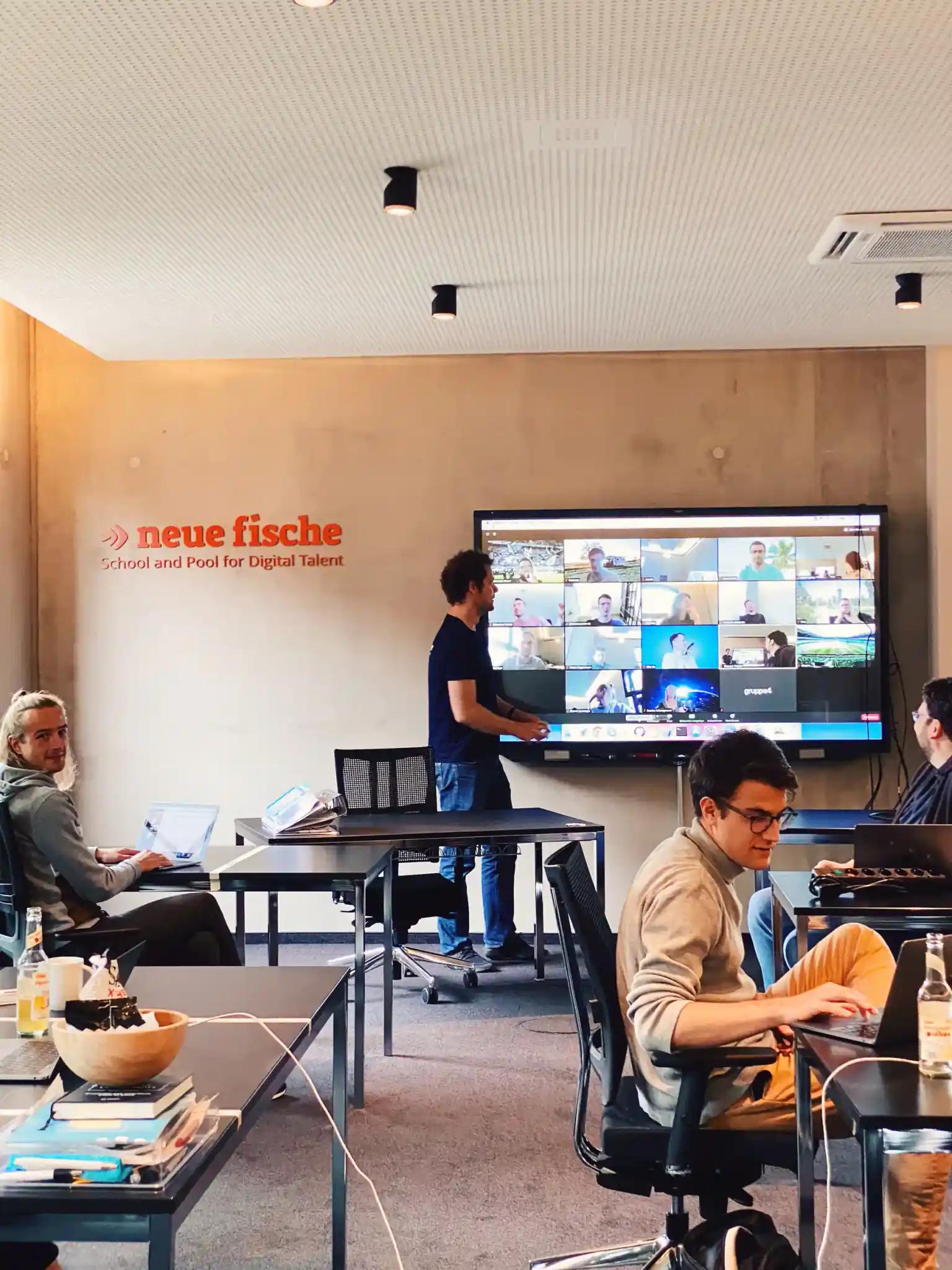 Moderner Schulungsraum mit Personen und Online-Meeting auf Bildschirm.