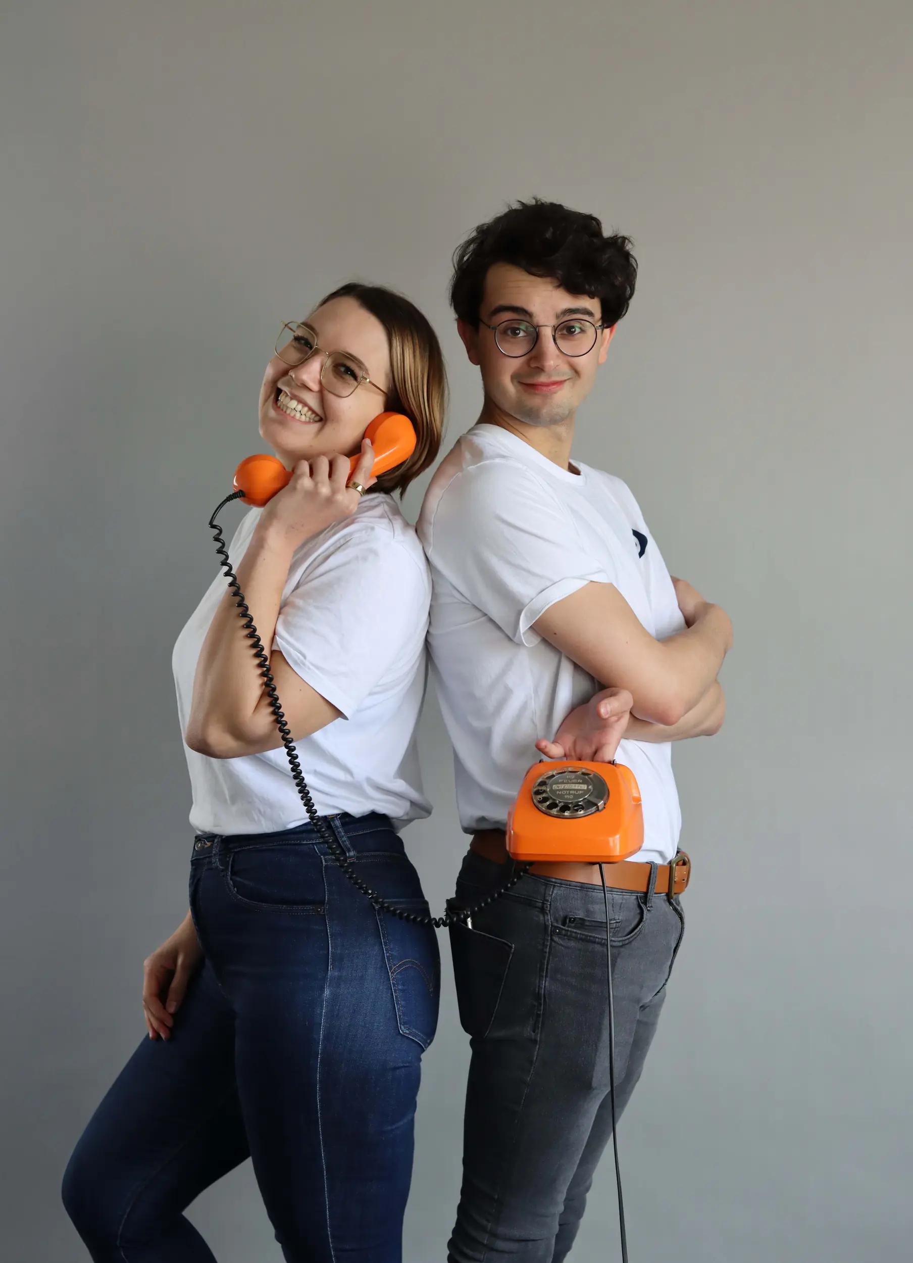 Two young adults standing back to back, one holding an orange retro telephone handset, the other holding the phone base.