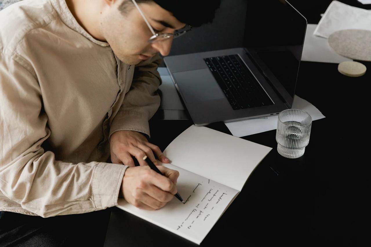 Person writing notes beside a laptop on a black table.