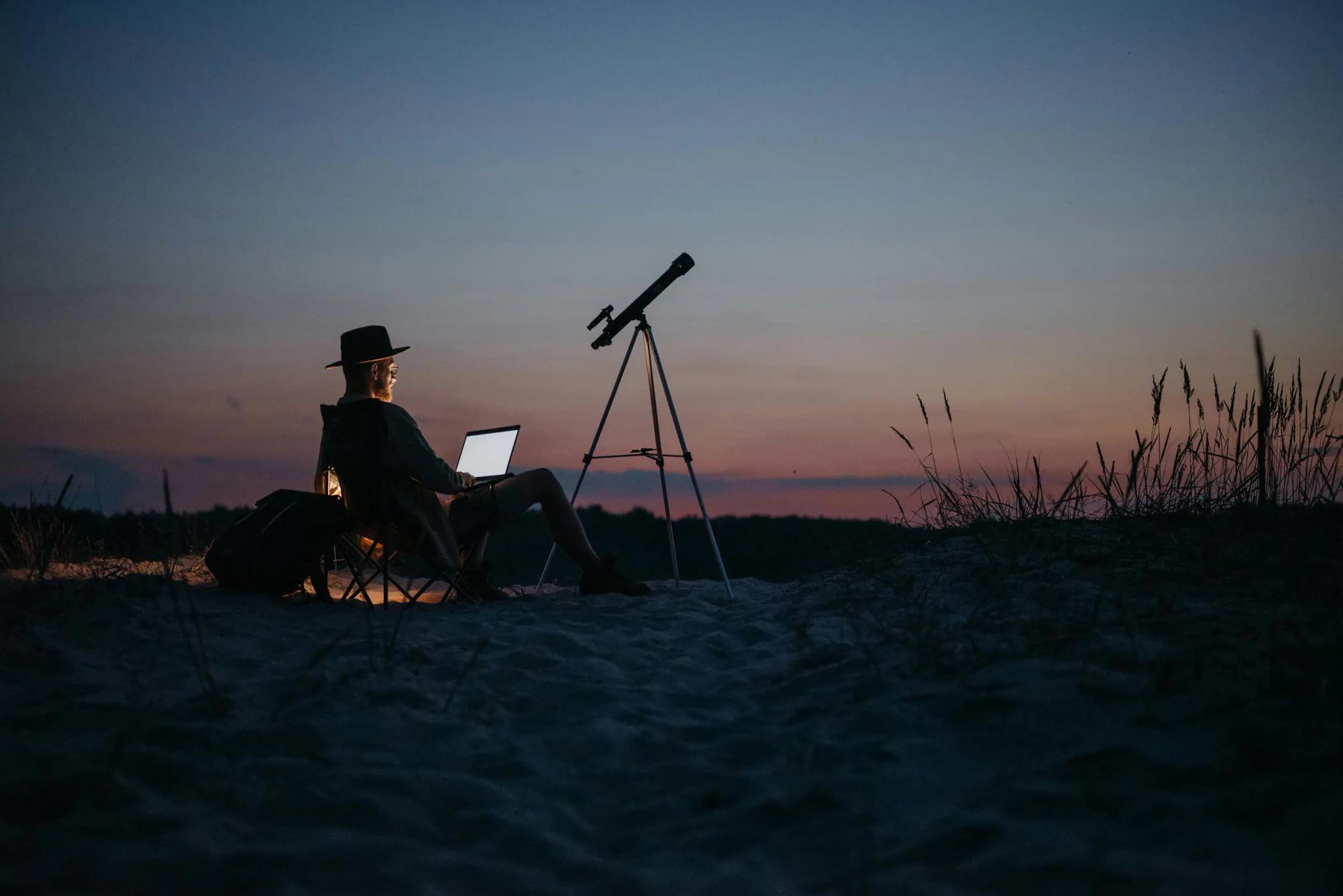 Person mit Laptop und Teleskop bei Sonnenuntergang im Freien