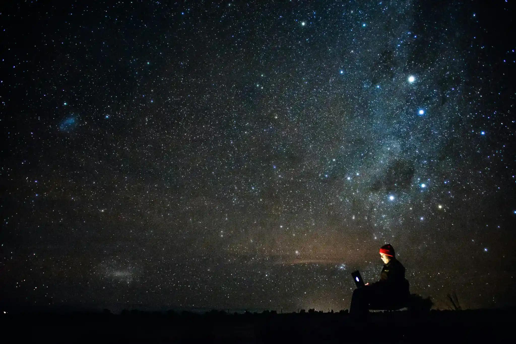 Person sitting under starry night sky with laptop.