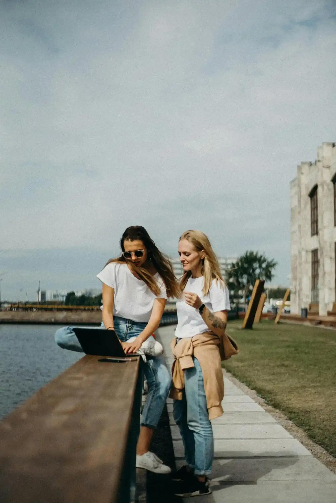 Two women with a laptop outdoors by the water.