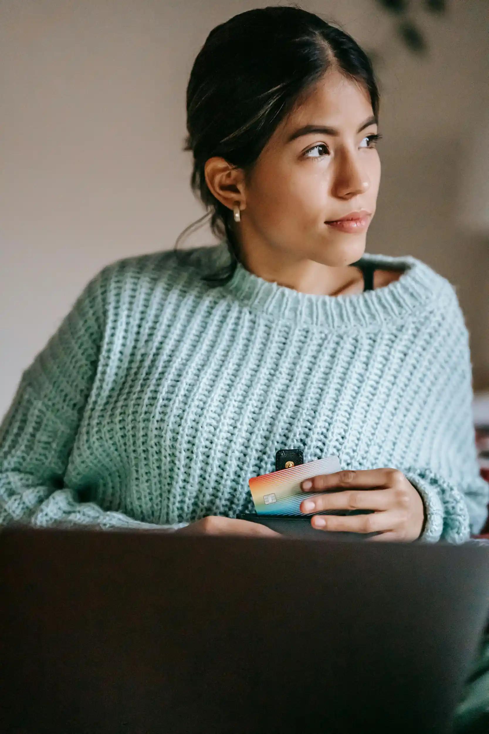 Pensive woman in sweater holding credit card in front of laptop.