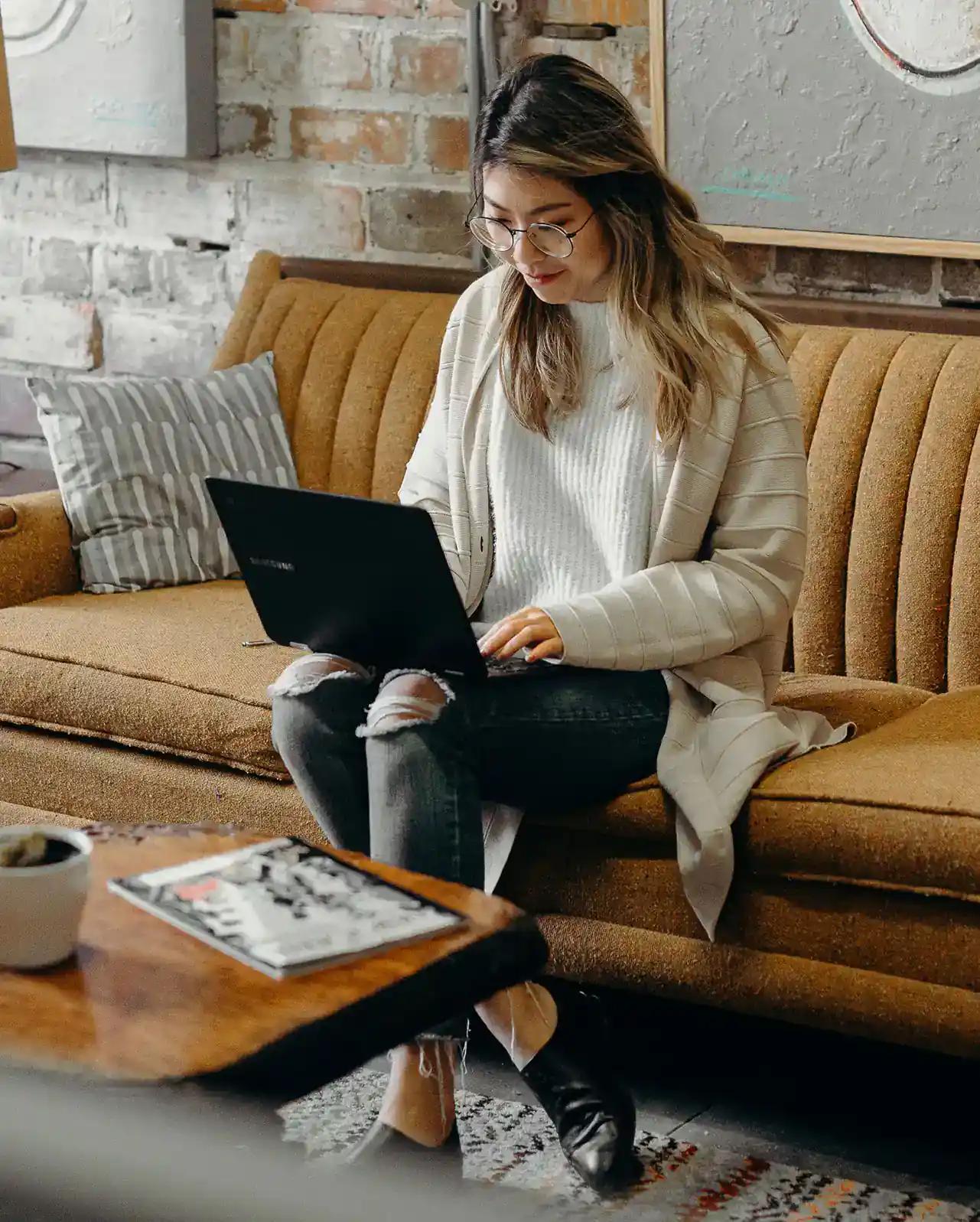 Woman working on a laptop sitting on a couch in a cozy room.