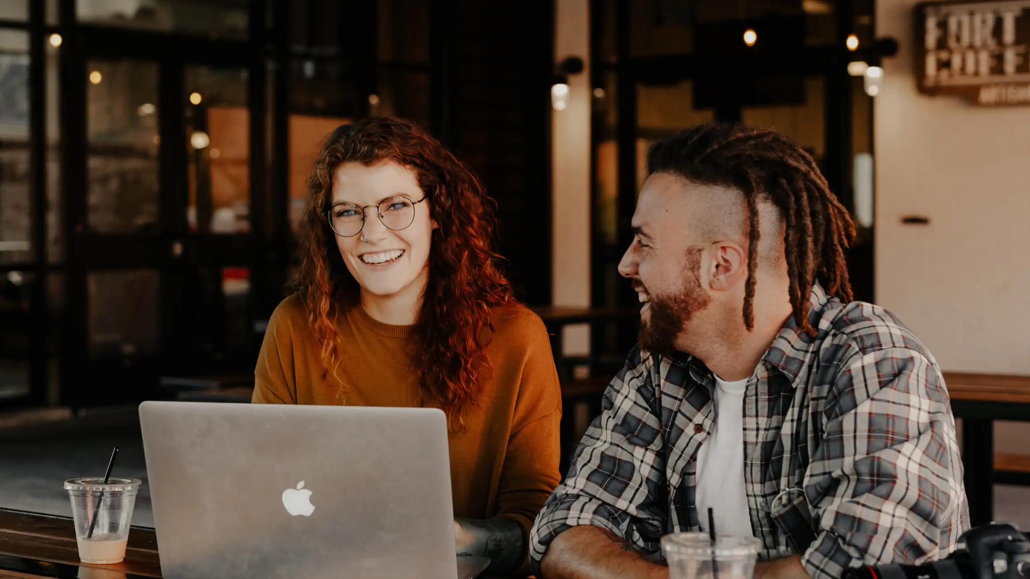 Zwei junge Erwachsene lächeln bei der Arbeit mit Laptop im Café.