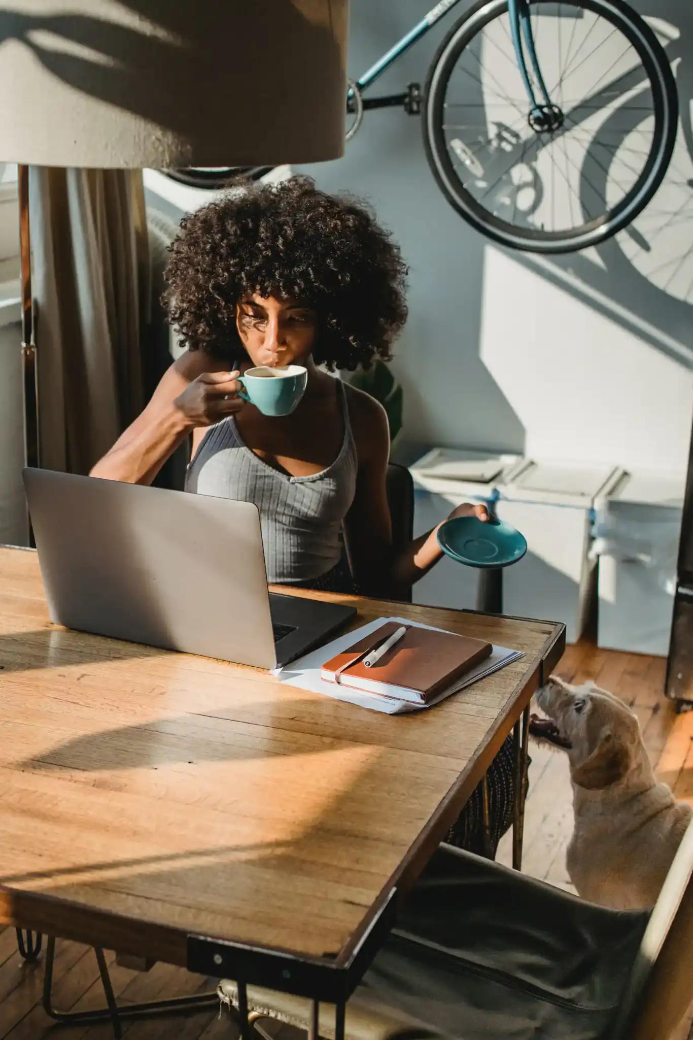 Woman drinking coffee at desk with laptop and dog nearby.