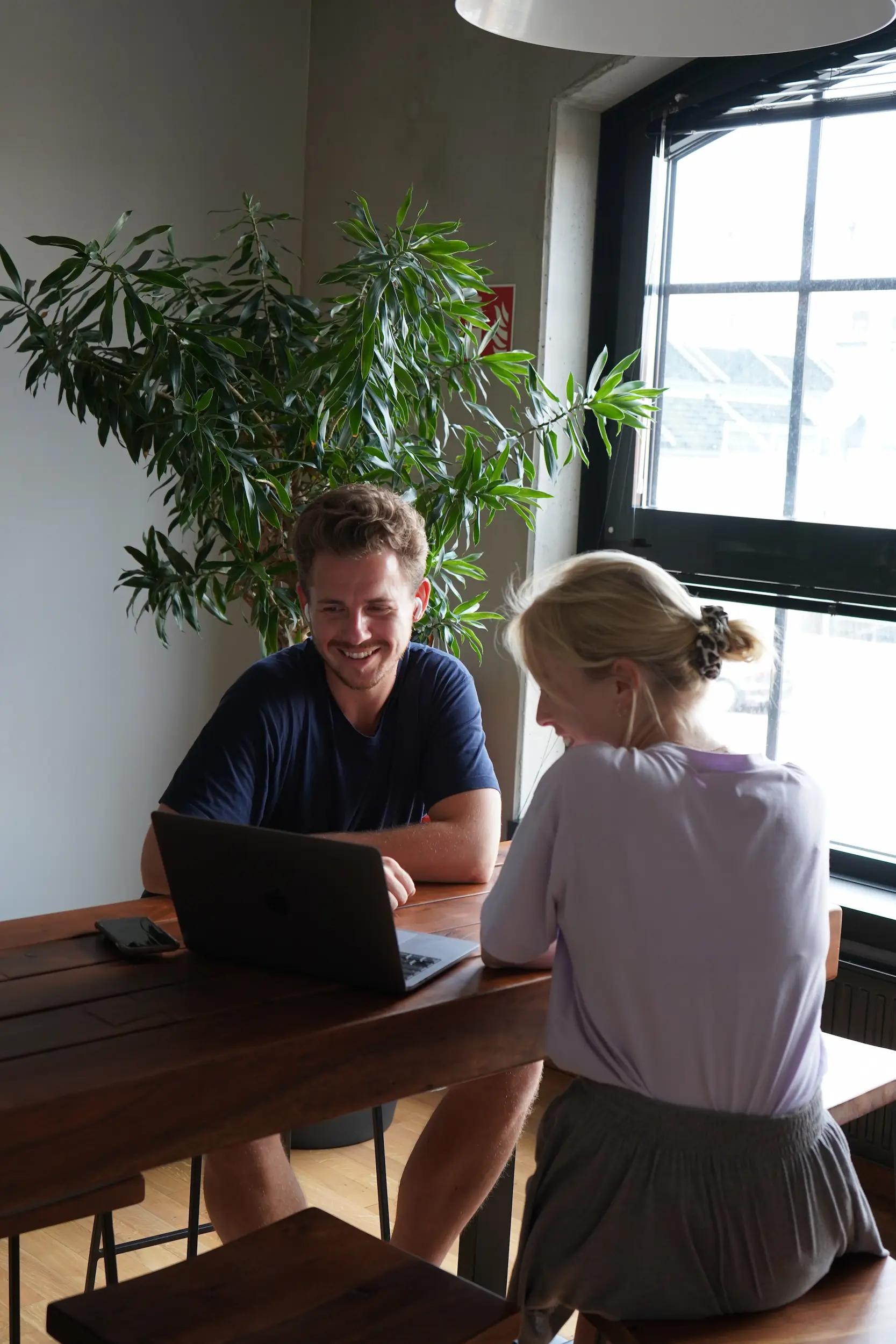 Zwei Personen arbeiten gemeinsam an einem Laptop an einem Holztisch.