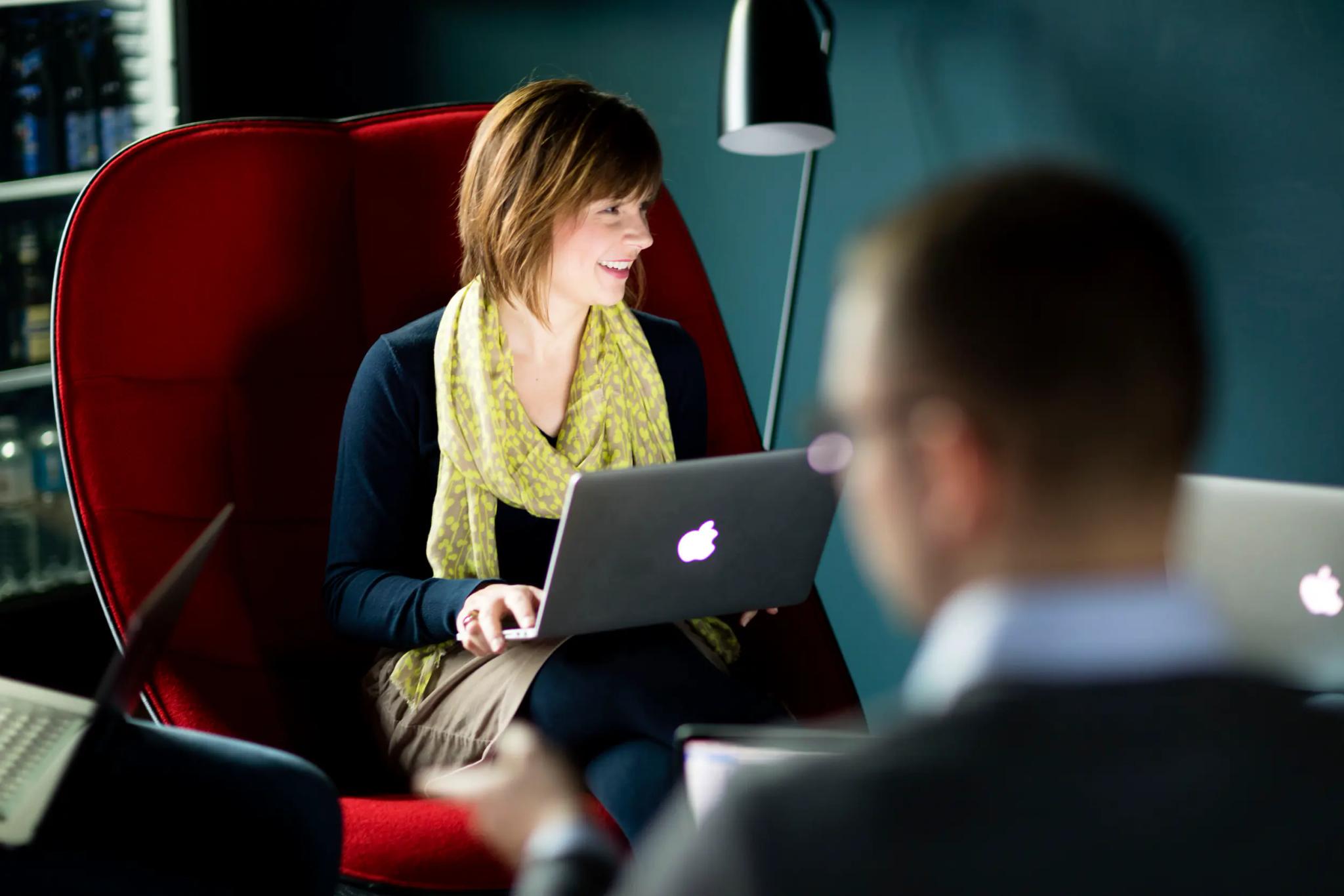 Frau sitzt mit Laptop in rotem Stuhl und spricht mit Kollegen.