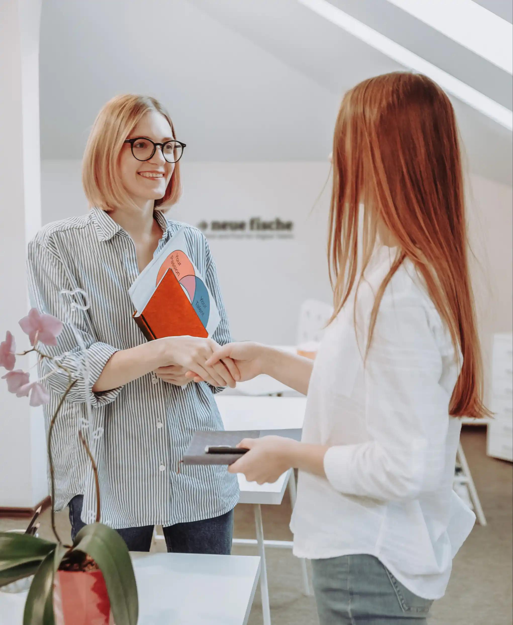 Two standing women shaking hands in an office space.